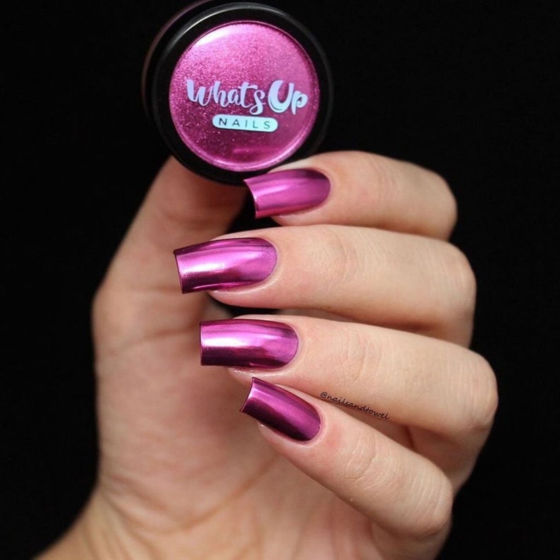 K&ouml;nnte beinhalten: Ein kleines Glas mit rosa metallischem Nagelpulver mit der Aufschrift "What's Up Nails". Eine Hand ist mit vier Fingern&auml;geln zu sehen, die mit einem gl&auml;nzenden, metallischen rosa Lack lackiert sind.