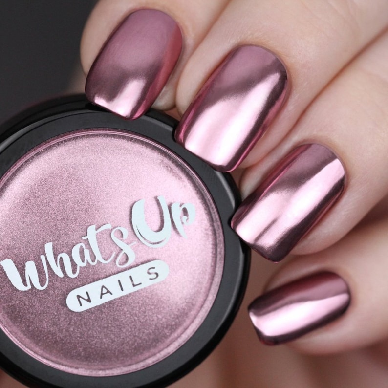May include: Close-up of a pink metallic nail polish with a shiny, chrome finish. The polish is in a small, round jar with a black lid and the text "What's Up Nails" printed on the label.