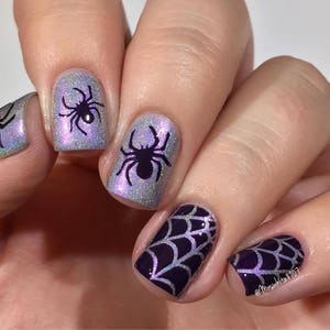 May include: Close-up of a hand with five fingernails painted with a purple and silver glitter polish. Each nail features a different design: a spider web, a spider, and a spider web. The nail art is a fun and festive way to celebrate Halloween.