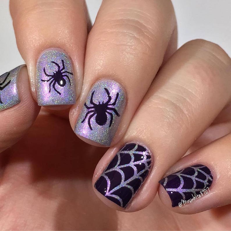 May include: Close-up of a hand with five fingernails painted with a purple and silver glitter polish. Each nail features a different design: a spider web, a spider, and a spider web. The nail art is a fun and festive way to celebrate Halloween.
