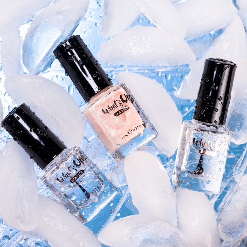 May include: Three clear glass bottles of nail polish on a blue background with ice cubes. The bottles are labeled "What's Up Nails" and contain a clear, pink, and white polish.