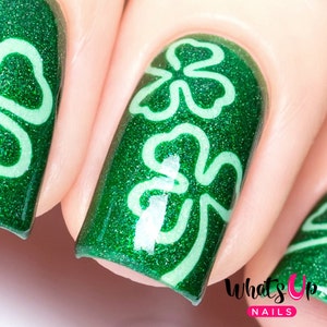 May include: Close-up of a green glitter nail polish with white shamrock designs. The nail polish is shiny and has a sparkly finish. The shamrocks are outlined in white and are a popular design for St. Patrick's Day.