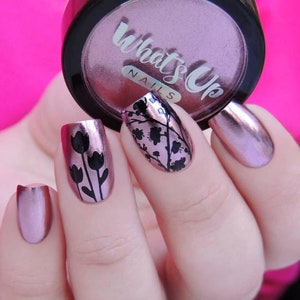 May include: Close-up of a hand with pink metallic nail polish. Two nails feature black floral designs, one with tulips and the other with a more abstract design. The image also shows a jar of "What's Up Nails" pink metallic nail polish.