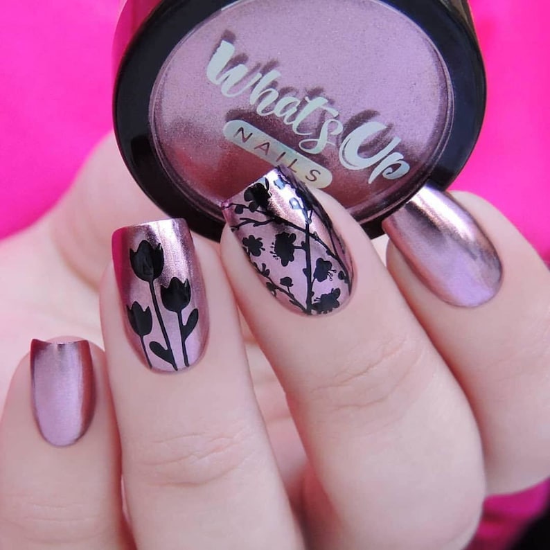 May include: Close-up of a hand with pink metallic nail polish. Two nails feature black floral designs, one with tulips and the other with a more abstract design. The image also shows a jar of "What's Up Nails" pink metallic nail polish.