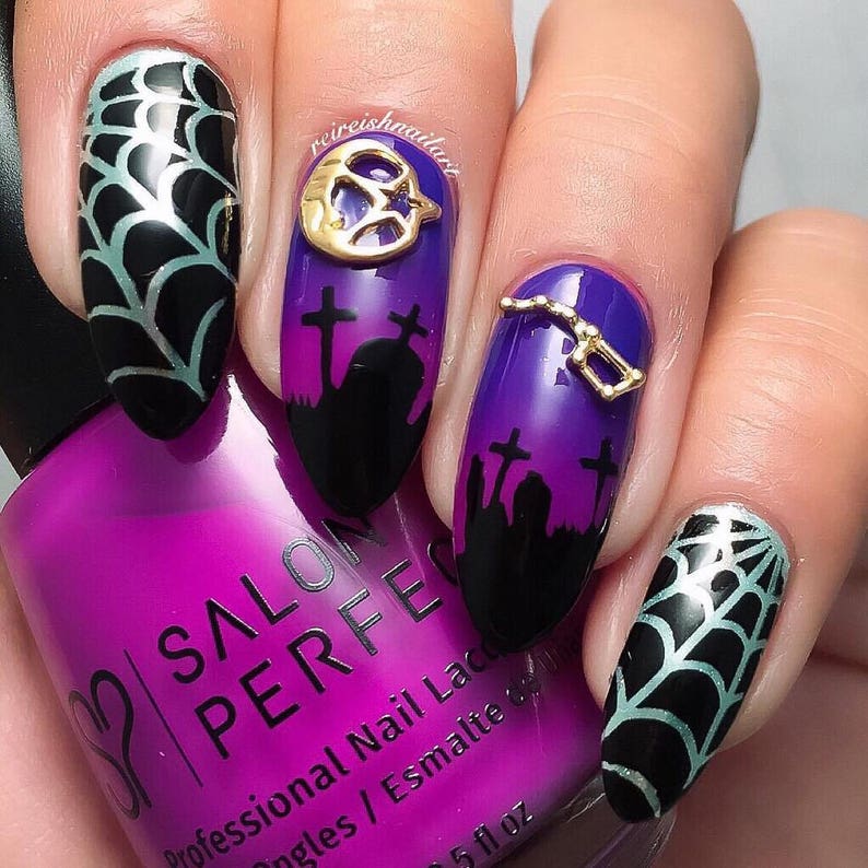 May include: Close-up of a hand with five nails painted with a Halloween theme. The nails are painted with a gradient of purple and black, with black silhouettes of tombstones and a spiderweb design. Two of the nails have gold charms, one shaped like a crescent moon and the other like a constellation. The hand is holding a bottle of purple nail polish labeled "Salon Perfect Professional Nail Lacquer."