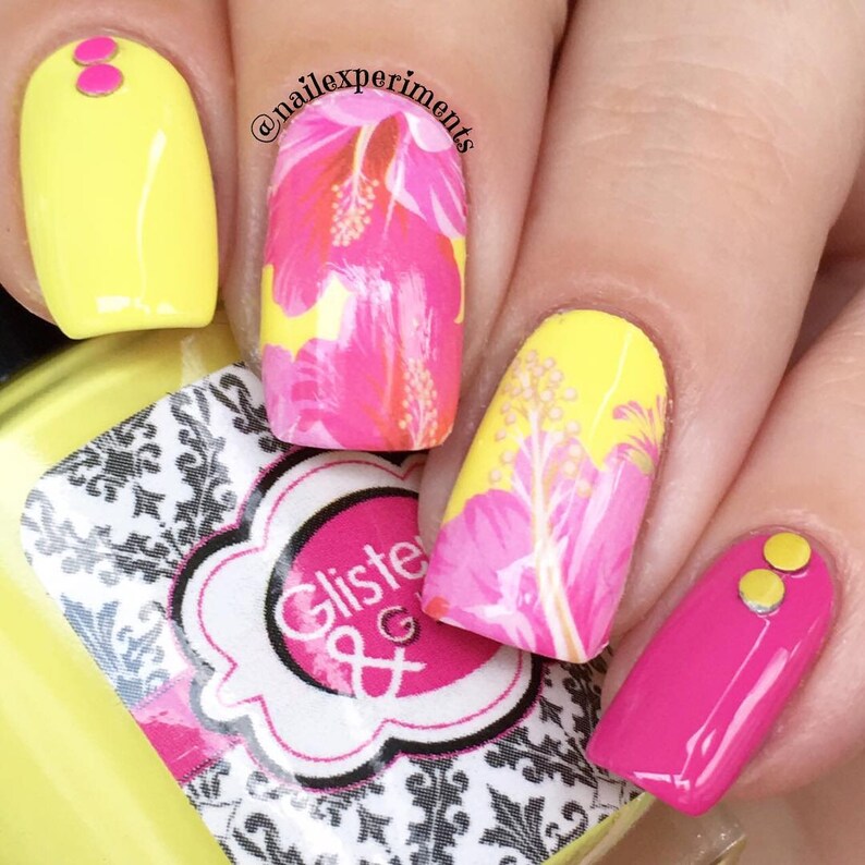 Puede incluir: Primer plano de u&ntilde;as pintadas con esmalte de u&ntilde;as amarillo y rosa. Las u&ntilde;as presentan un dise&ntilde;o floral con flores de hibisco rosas y hojas verdes. Dos u&ntilde;as tienen puntos dorados encima del esmalte.