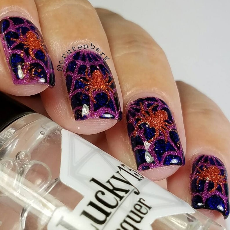 May include: Close-up of a hand with four fingernails painted with a dark purple and orange spiderweb design. The nail polish is glittery and the spiderwebs are outlined in orange. The hand is holding a bottle of clear nail polish with the text "Lucky 13" and "Top Coat" on the label.