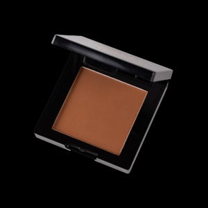 May include: A square, open compact of bronzer with a matte finish. The bronzer is a warm, medium brown shade, set in a black and white plastic case. The compact is angled against a black background.