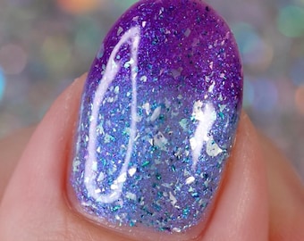 May include: Close-up of a fingernail with a purple and blue gradient nail polish. The polish has a glittery finish with iridescent flakes.
