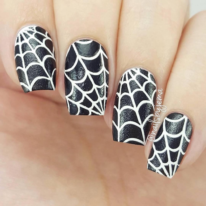 May include: Close-up of four fingernails painted black with white spiderweb designs. The nails are short and square shaped. The design is intricate and detailed, with thin white lines creating a web-like pattern.