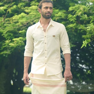 May include: A cream-coloured button-down shirt with a small embroidered elephant logo on the chest. The shirt is paired with a matching cream-coloured wrap skirt, featuring a thin red and cream stripe detail. The outfit is worn outdoors.