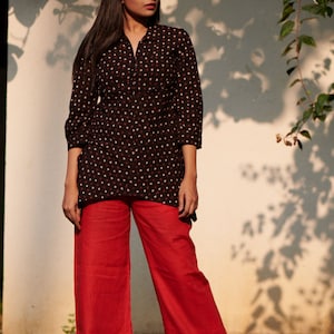 May include: A person wearing a black tunic with a white and red triangle pattern, paired with bright red wide-leg pants. The outfit is styled in a natural outdoor setting, with sunlight and shadows.