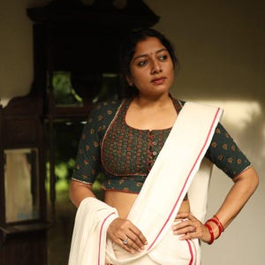 May include: A woman wearing a traditional Indian outfit. The ensemble includes a dark green blouse with a red and brown pattern, paired with a white saree featuring red trim. The woman is also wearing red bangles.