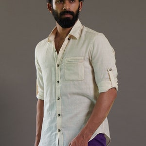 May include: A man wearing a light beige linen shirt with a button-down collar and a single chest pocket. He is also wearing a purple dhoti.
