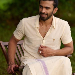 May include: A cream-coloured short-sleeved shirt with a collar and a small palm tree emblem. The shirt is worn with a matching sarong-style wrap with lavender stripes. The person is seated on a wooden chair.