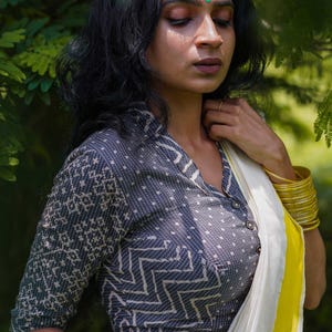 Puede incluir: Una blusa gris oscuro con un patrón geométrico y cierre de botones. La blusa tiene mangas tres cuartos y se combina con un sari blanco y amarillo. Brazaletes dorados adornan la muñeca.