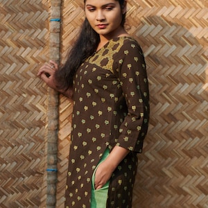 Puede incluir: Una túnica marrón con un diseño floral amarillo en el escote y pequeños estampados florales amarillos. La túnica se combina con pantalones verde claro. El fondo es una pared de bambú tejido.