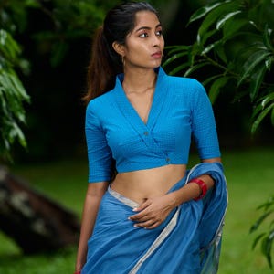 May include: A blue, short-sleeved blouse with a wrap-style front and a high collar. The blouse is paired with a matching blue saree. The image is set against a backdrop of green foliage.