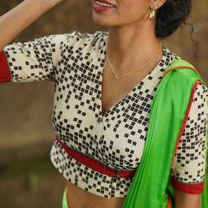 May include: A white and black geometric print blouse with a red and green trim. The blouse has a button closure and a fitted style. The model is wearing a green saree.