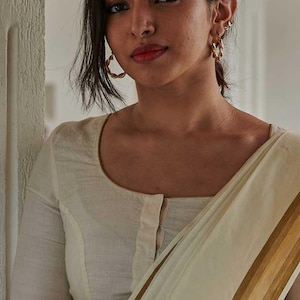 May include: A woman wearing a cream-colored blouse with a scoop neckline and a matching saree. The saree has gold stripes. She is wearing gold hoop earrings and has a bindi on her forehead.