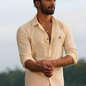 May include: A cream-coloured button-down shirt with long sleeves. The shirt features a small embroidered design on the chest. The person is also wearing a gold necklace and red trousers. The background is a blurred outdoor scene.