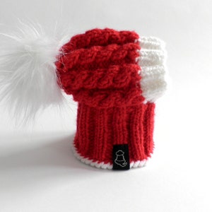 May include: Red and white knitted beanie with a white pom pom. The beanie has a cable knit pattern and a white trim.