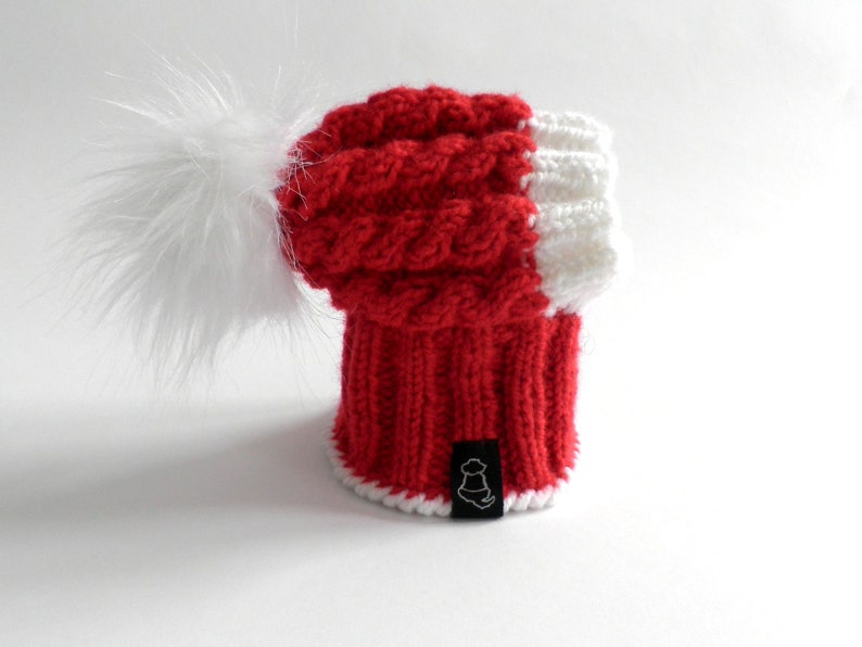 May include: Red and white knitted beanie with a white pom pom. The beanie has a cable knit pattern and a white trim.