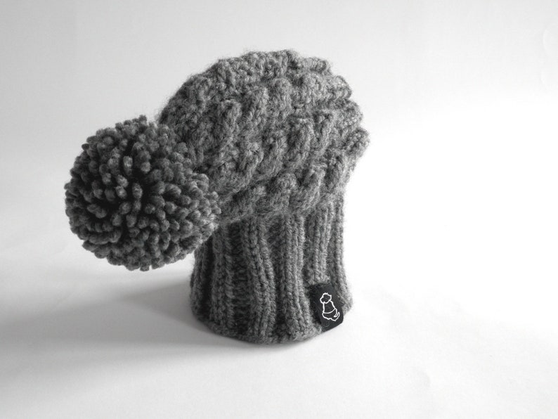 May include: Gray knitted beanie with a large pom pom on top. The beanie has a cable knit pattern and a small black label with a white logo.