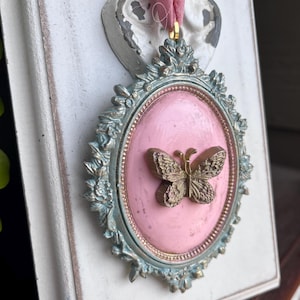 May include: A pink oval frame with a gold butterfly design. The frame is decorated with ornate details and has a light blue finish.