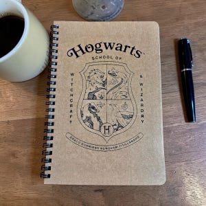 May include: A spiral-bound notebook with a brown cover featuring the Hogwarts School of Witchcraft & Wizardry crest. The crest includes a lion, snake, badger, and eagle. A black pen and a mug of coffee are also visible.