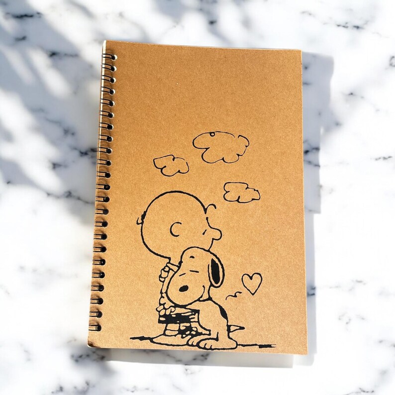 May include: A brown spiral notebook with a black and white illustration of Snoopy and Charlie Brown hugging. The illustration includes a heart and clouds.