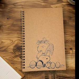 May include: A brown spiral bound notebook with a Peanuts cartoon of Charlie Brown and Snoopy on the cover. Snoopy is holding a surfboard and Charlie Brown is looking up at the sky.