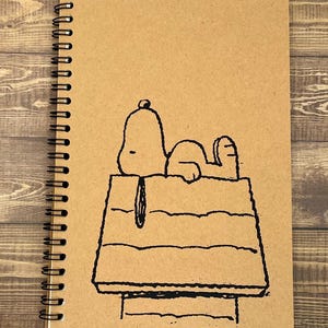 May include: A brown notebook with a black spiral binding. The cover features a black and white line drawing of Snoopy sleeping in his doghouse.