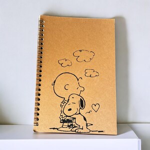 May include: A brown spiral notebook with a black and white illustration of Snoopy and Woodstock. The illustration shows Snoopy hugging Woodstock with a heart above them.