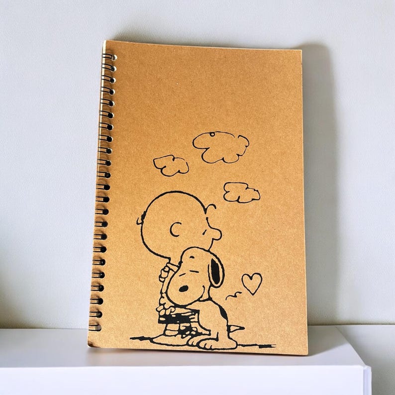May include: A brown spiral notebook with a black and white illustration of Snoopy and Woodstock. The illustration shows Snoopy hugging Woodstock with a heart above them.