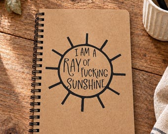May include: A spiral-bound notebook with a tan kraft paper cover featuring a black sun graphic and the text I AM A RAY OF FUCKING SUNSHINE printed in the center. The notebook has a black metal coil binding along the left side of the cover.