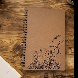 May include: Spiral-bound notebook with a brown cover featuring a black illustration of Snoopy jumping into a pile of leaves. The notebook is on a wooden surface, with a second notebook and a coffee cup visible.