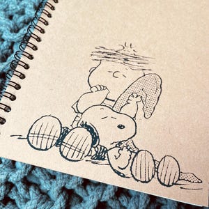 May include: A brown spiral notebook with a black and white illustration of Snoopy and Woodstock. The illustration shows Snoopy lying on his back with Woodstock perched on his stomach.