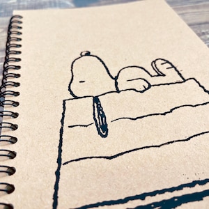 May include: A brown spiral notebook with a black and white illustration of Snoopy lying on his doghouse.