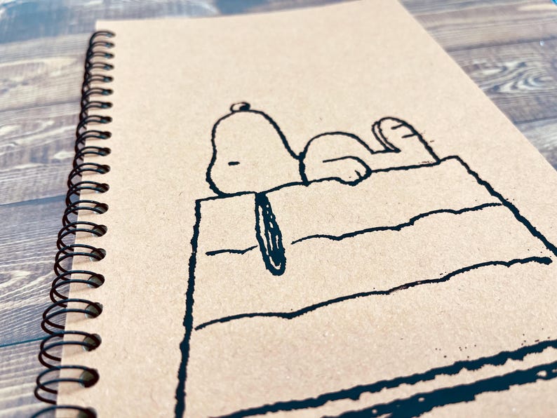 May include: A brown spiral notebook with a black and white illustration of Snoopy lying on his doghouse.