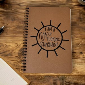 May include: A spiral-bound notebook with a brown cover. The cover features a black sun design with the text "I AM A RAY of FUCKING SUNSHINE". The notebook is on a wooden surface, with a pen and another notebook visible.