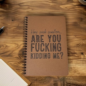 May include: A spiral-bound notebook with a brown cover and the text "Hey quick question... ARE YOU FUCKING KIDDING ME?" in black. The notebook is on a wooden surface, with a pen and a small open notebook visible.