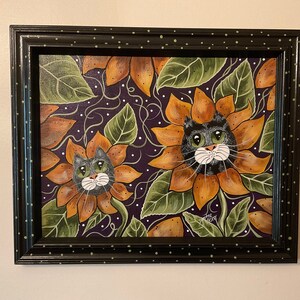 May include: A framed painting of two black cats with white faces peeking out of sunflowers. The sunflowers are orange and yellow with green leaves. The background is a dark purple with white dots.