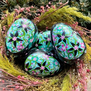 May include: Four painted rocks in a nest of dried grasses. The rocks are painted with a floral design in shades of black, green, pink, and turquoise.
