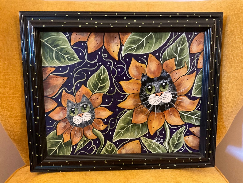 May include: Framed painting of two black cats with green eyes peeking out of sunflowers on a purple background with white dots.