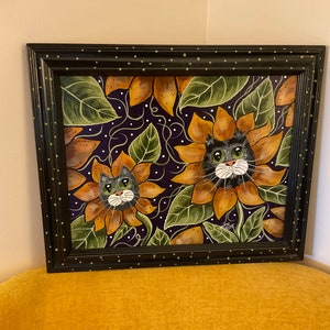 May include: A framed painting of two cats peeking out of sunflowers. The sunflowers are yellow and orange with green leaves. The background is a dark purple with white dots.