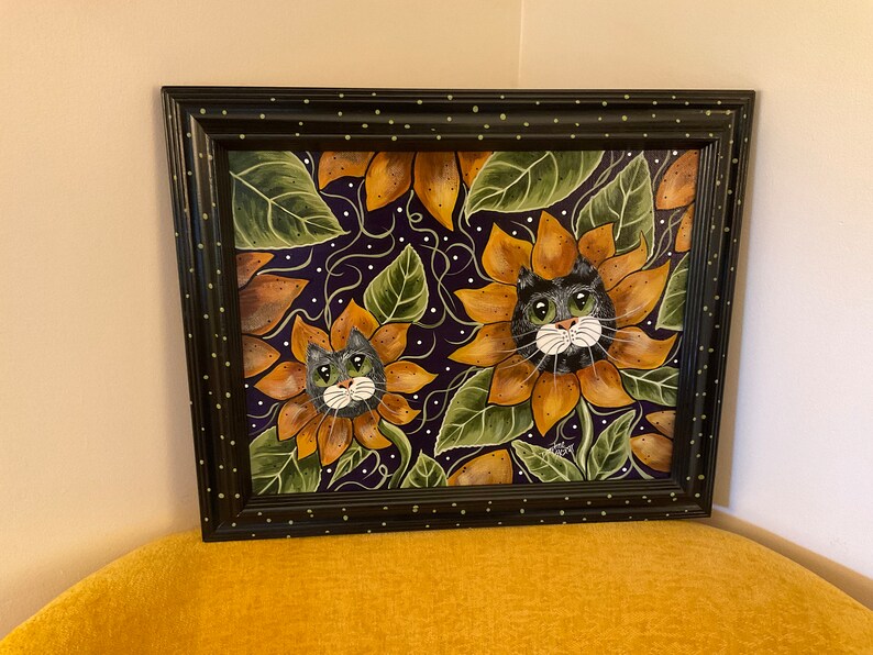 May include: A framed painting of two cats peeking out of sunflowers. The sunflowers are yellow and orange with green leaves. The background is a dark purple with white dots.