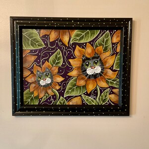 May include: Framed painting of two cats with green eyes and white whiskers, peeking out of sunflowers on a purple background.