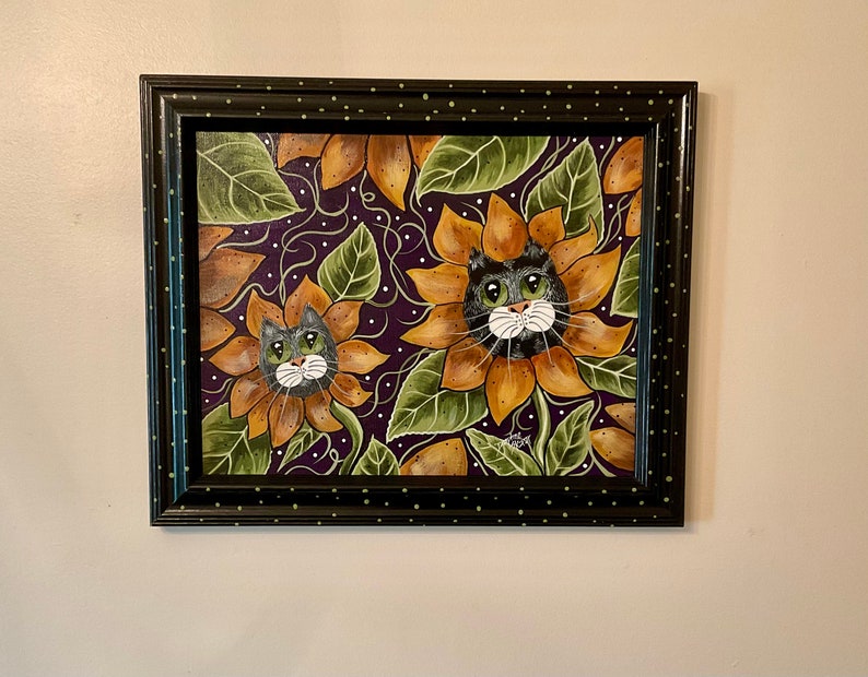 May include: Framed painting of two cats with green eyes and white whiskers, peeking out of sunflowers on a purple background.