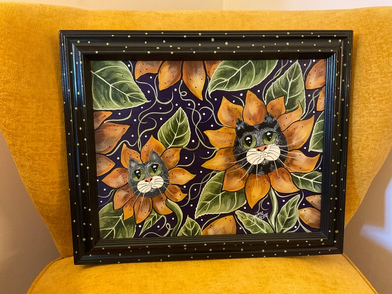 May include: Framed painting of two black cats with green eyes peeking out of sunflowers on a purple background with white dots.
